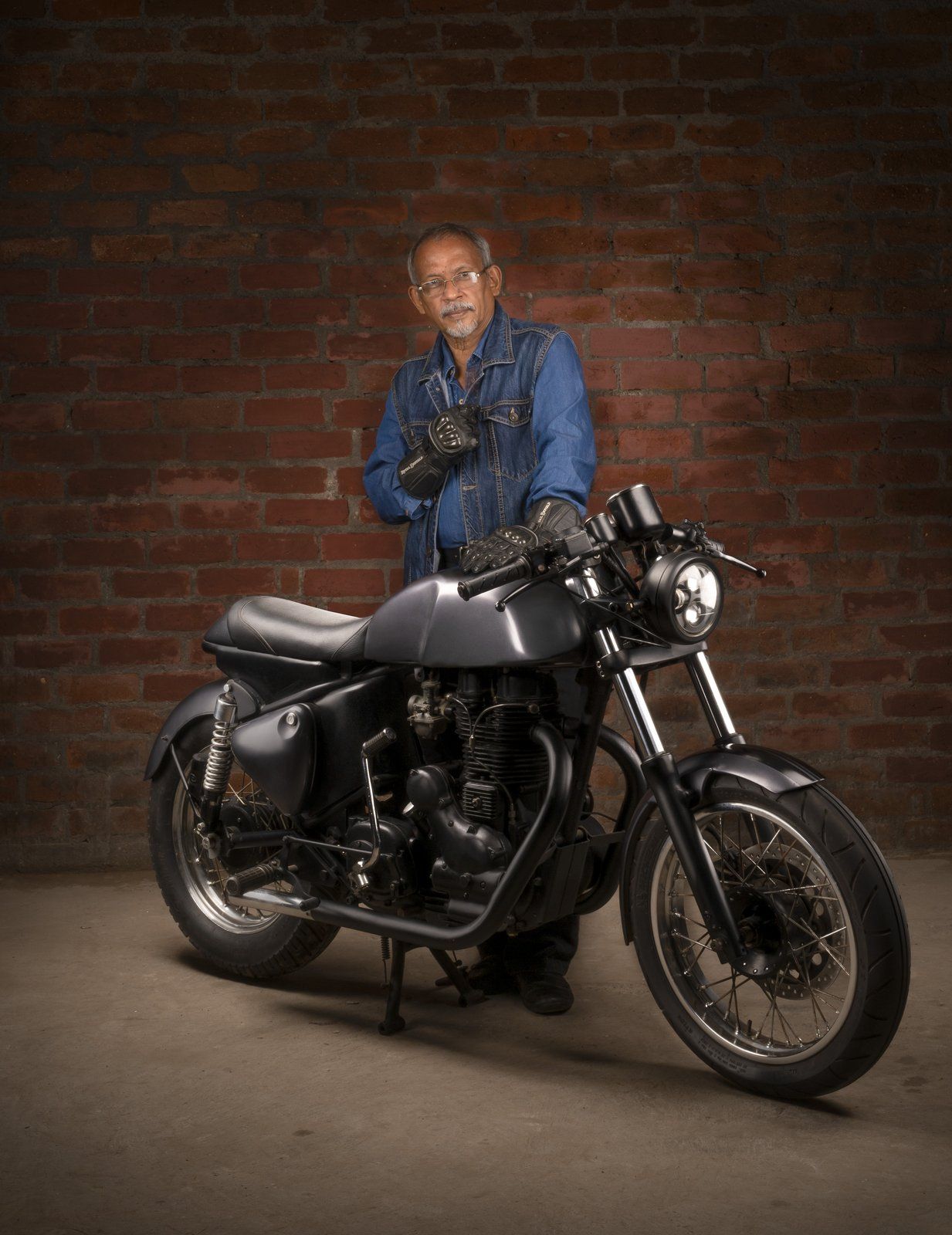 Man in a denim jacket standing with his black cafe racer motorcycle against a brick wall